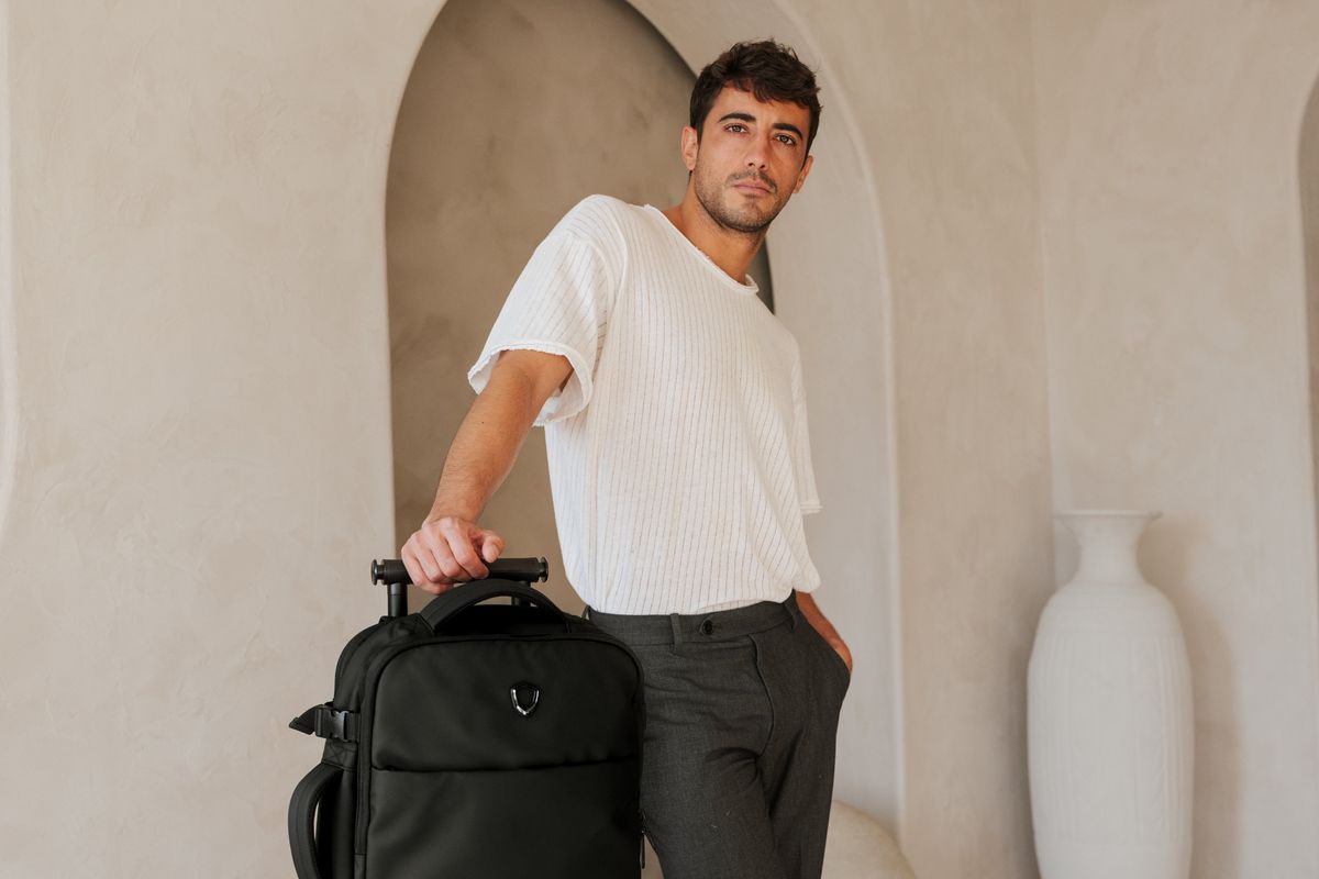 Man holding the telescoping handle of a black soft-sided Traveler's Choice wheeled travel bag in a sunlit archway