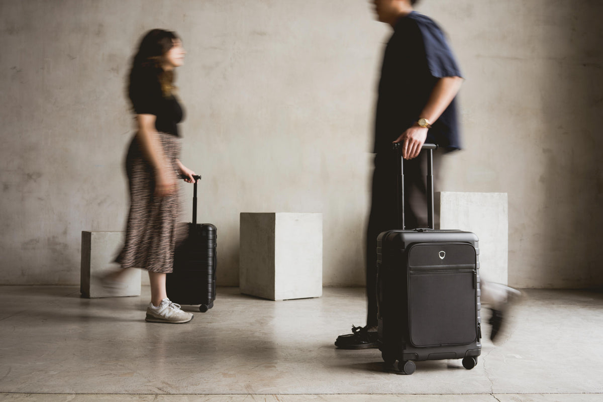 Two travelers walking with black Millennial front pocket carry-ons in a concrete hallway