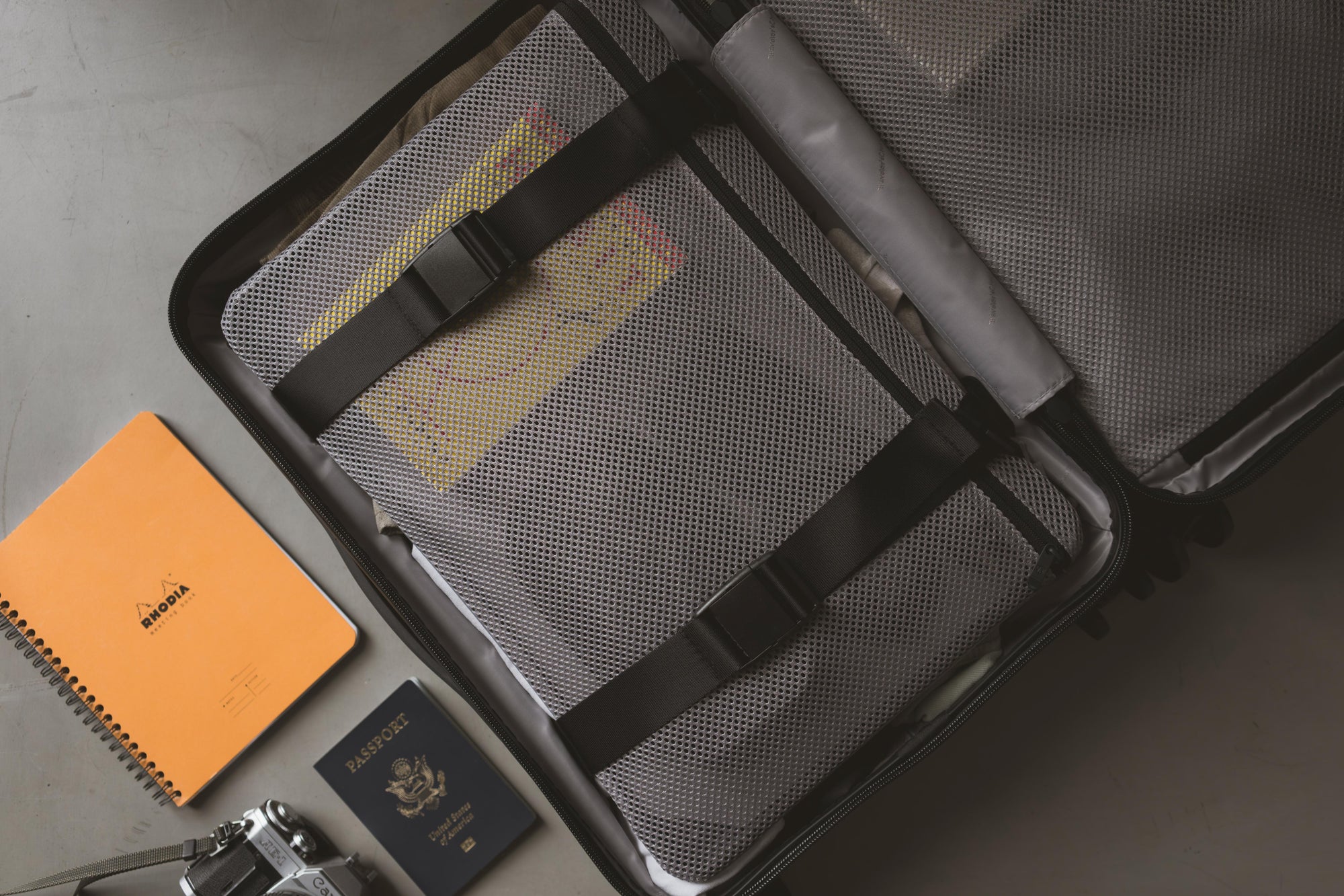 Inside of a Millennial carry-on packed under its mesh compression panel, with a passport and notebook alongside