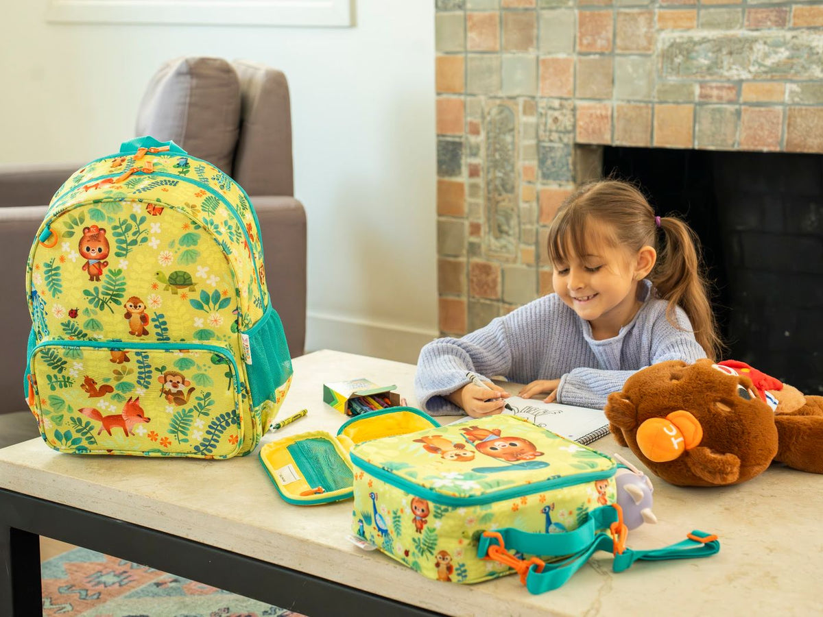 A girl draws at a table next to the Jungle backpack, lunch bag and pencil bag.