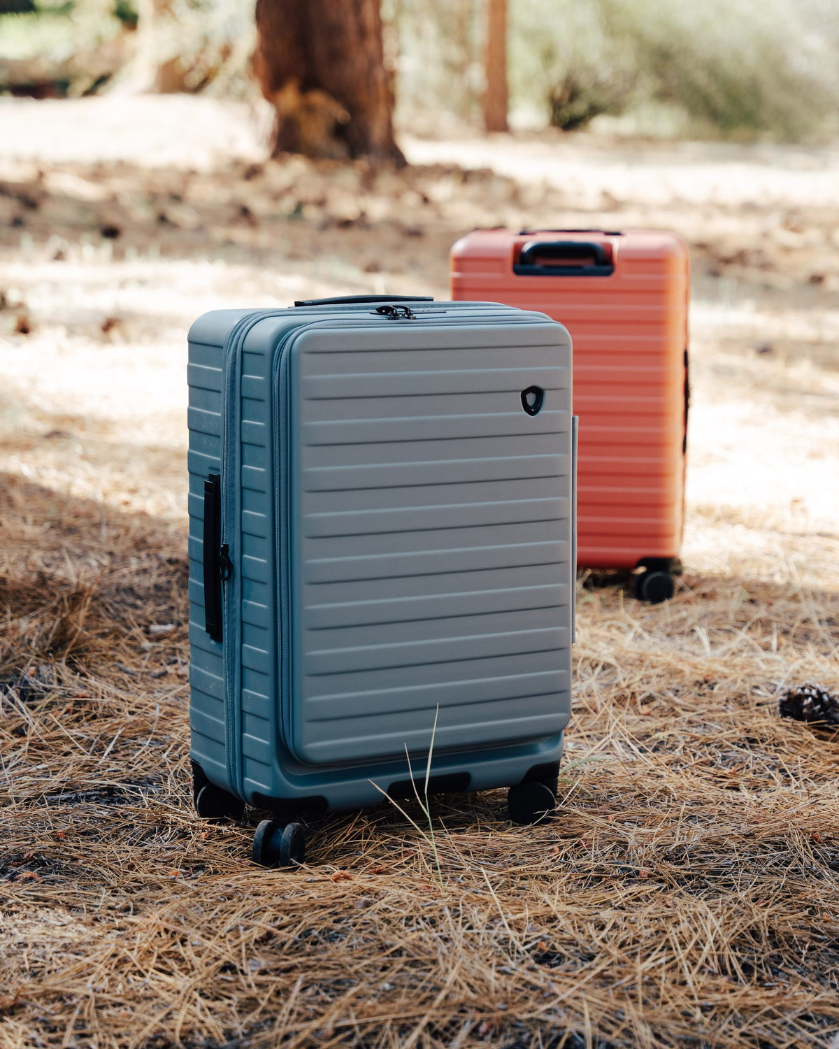 Slate blue Novia front-pocket carry-on standing on pine needles with an orange Novia suitcase behind it