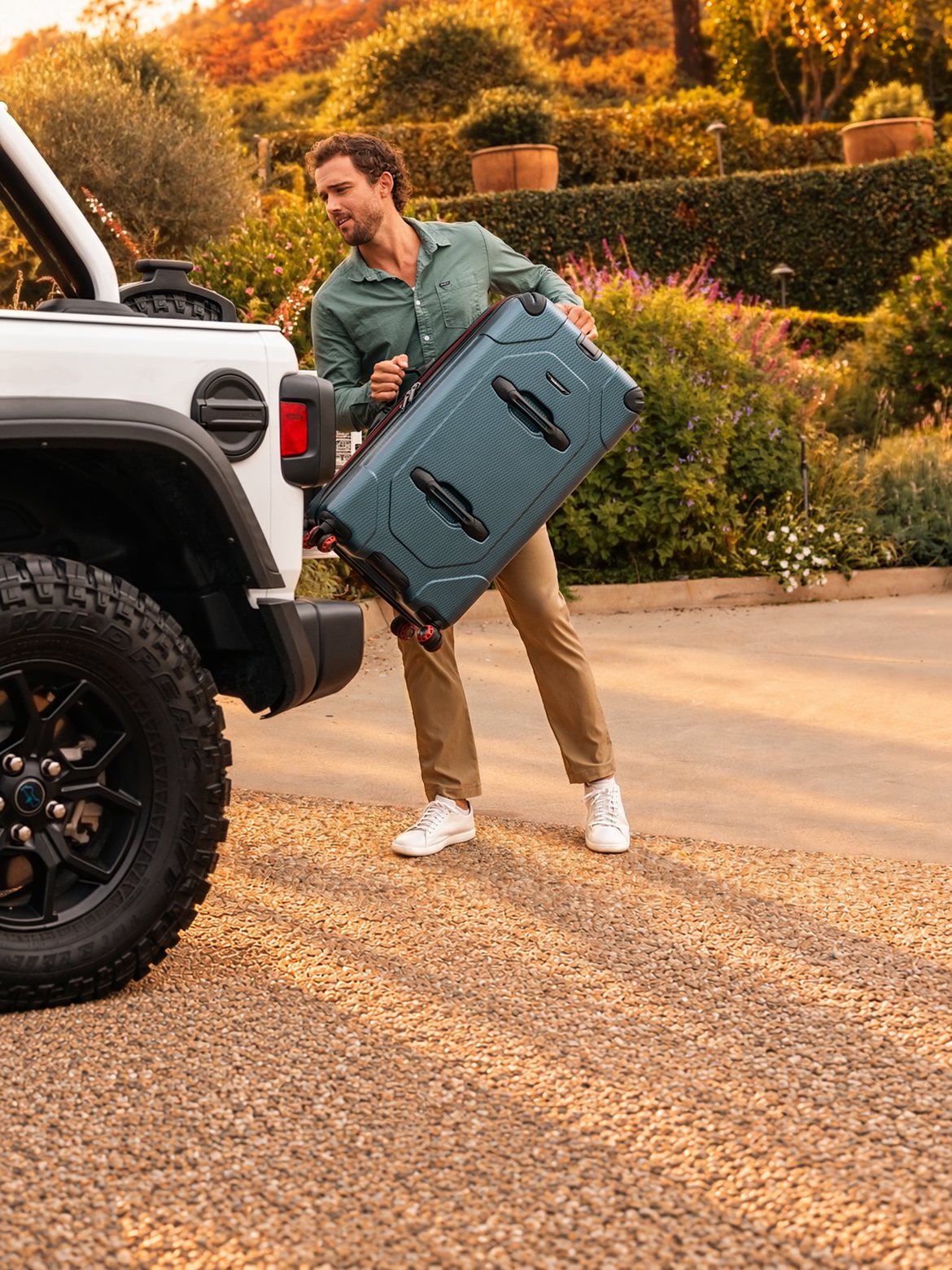 A man lifts a teal Traveler's Choice suitcase into a white Jeep outside a house with an autumn wreath on the front door.