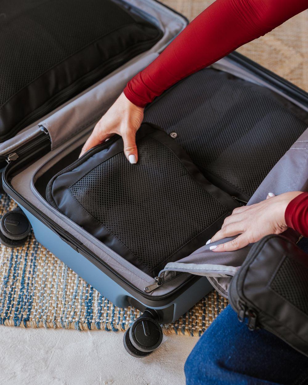 Two hands lowering a black mesh-front packing cube into an open light blue carry-on