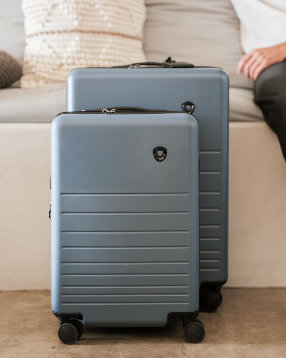 A slate blue carry-on standing in front of a larger matching check-in case beside a low upholstered daybed