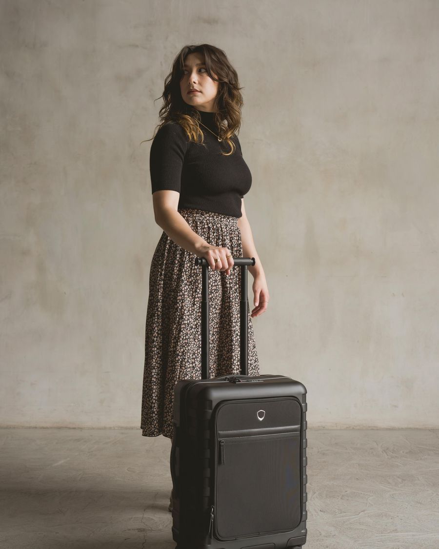 A woman against a warm plaster wall resting one hand on the extended handle of a black front-pocket carry-on