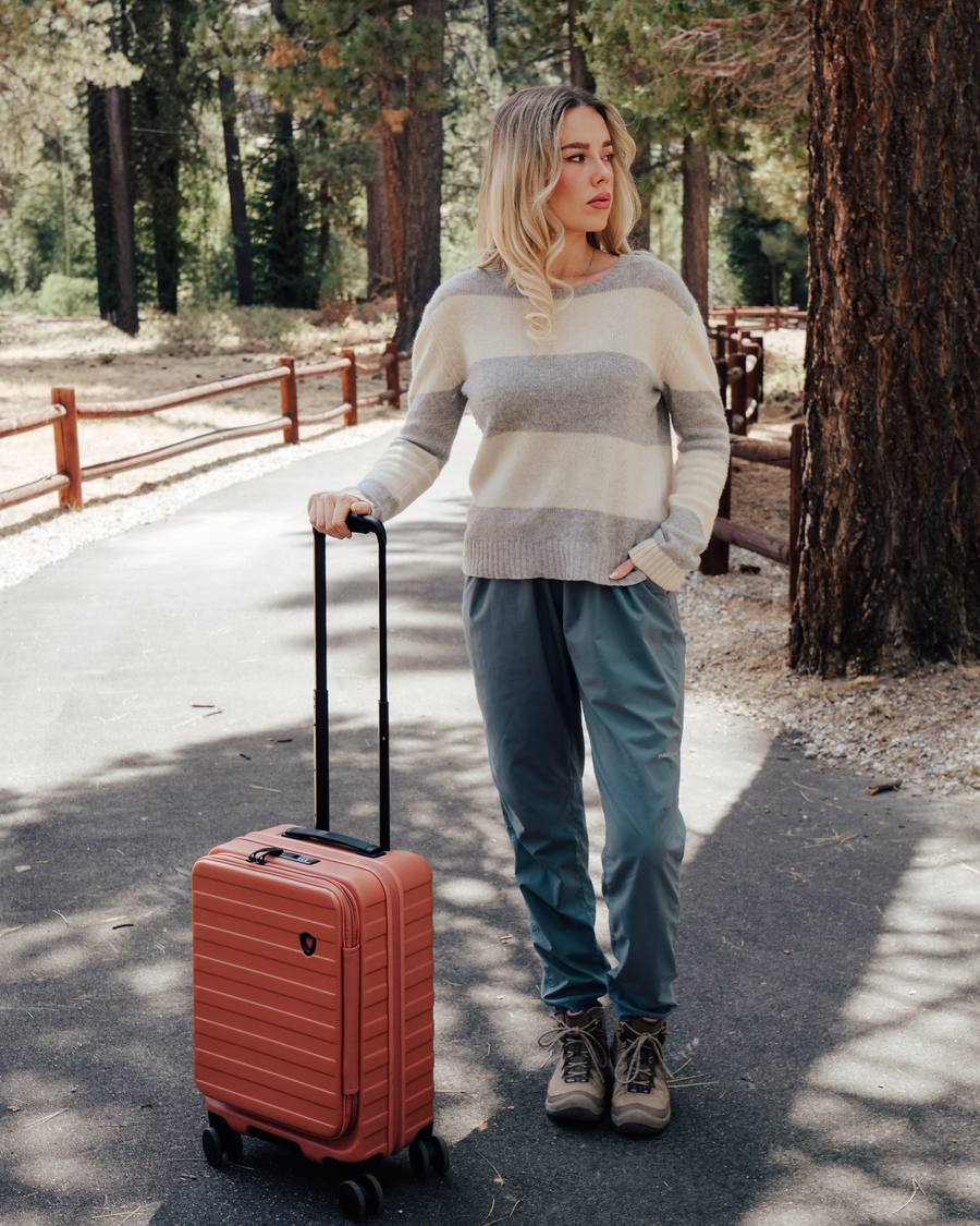 A woman in a striped sweater stands on a wooded path with one hand on the extended handle of a terracotta hard-shell carry-on