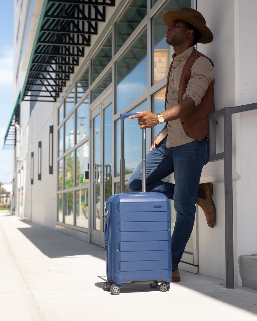 A man in a suede vest and hat leans against a storefront window, a slate blue hard-shell carry-on beside him with its handle extended